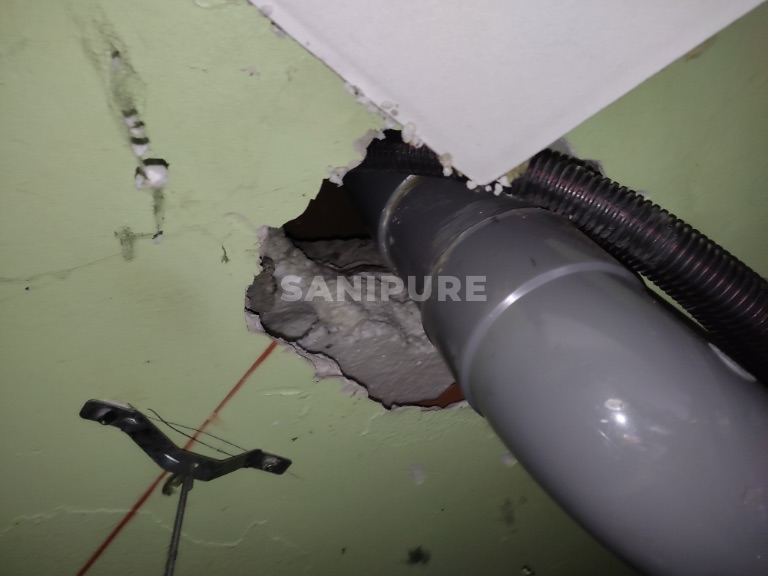 Tuyau d'évacuation gris et gaine noire traversant un mur vert par un trou aux bords cassés