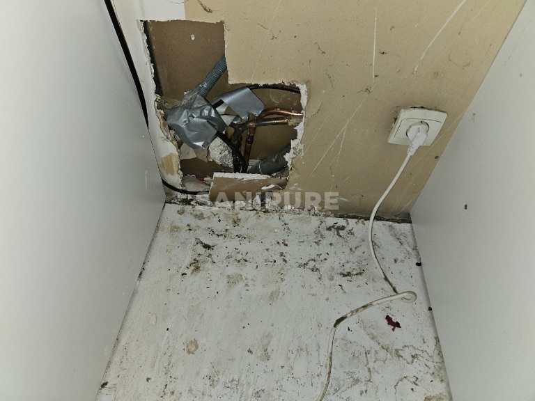 Fond de placard percé d'une ouverture autour de gaines techniques, plancher blanc souillé de crottes et débris
